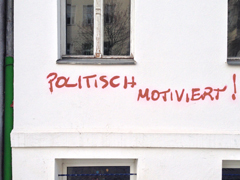 Foto zeigt weiße Hauswand. Auf dieser steht in rot gesprüht: politisch motiviert!